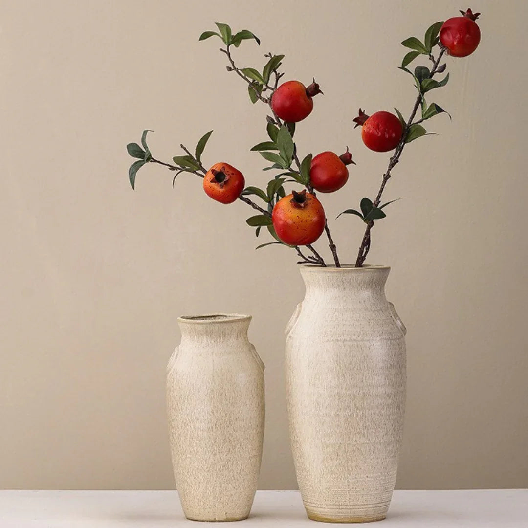 Two beige ceramic vases with branches of red pomegranate fruit and green leaves inside.