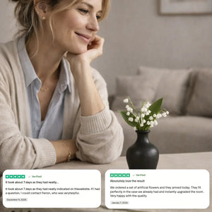 Smiling woman seated on sofa admiring artificial white flowers in a black vase with customer reviews displayed.